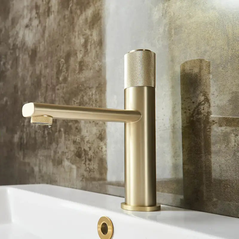 A modern, cylindrical Knob Switch Solid Brass Bathroom Sink Faucet in brushed gold by Wanfan installed on a white countertop. The faucet features a single lever and minimalist design, accented with textured detailing on the handle for improved grip. In the background, part of the rectangular sink and additional bathroom faucets can be seen.