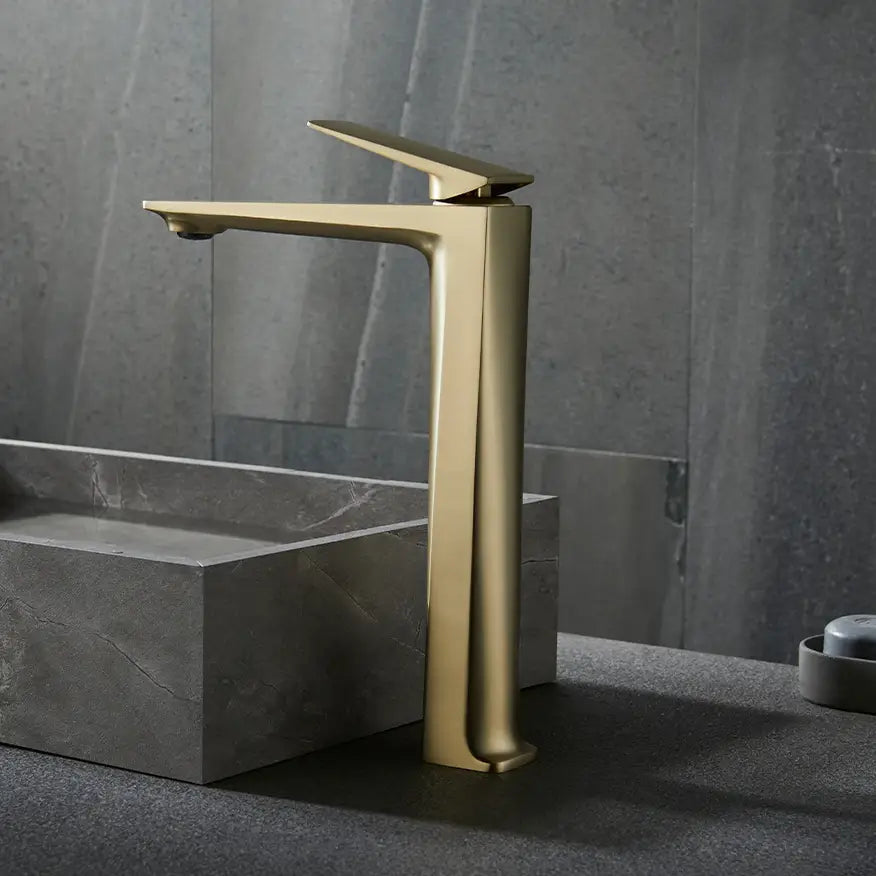 A modern, gold-colored Bagnolux Single Handle Brass Bathroom Sink Faucet with a sleek design is mounted on a grey stone countertop, pouring water into a matching rectangular grey stone sink. The backdrop features a textured grey wall, completing this elegant and cohesive look.