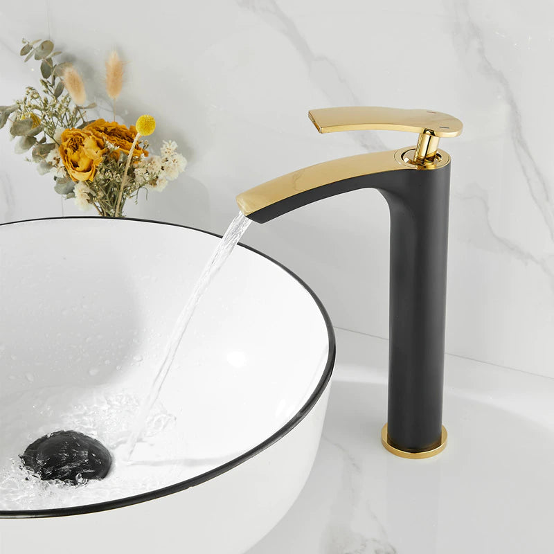 A modern bathroom sink with an allfixture Single Handle Bathroom Basin Faucet in white and gold. Water is flowing from the faucet into a round white basin with a black rim. A small bouquet of yellow and dried flowers rests on the marble countertop beside the sink.