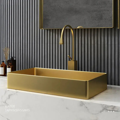 A modern bathroom showcasing the SUS304 Stainless Steel Rectangle Bathroom Sink in Brushed Gold by Mike Jake, set on a white marble countertop. Above the sink is a matching gold rectangular faucet. The wall behind features vertical gray stripes and a large rectangular mirror. To the left of the sink, two brown bottles and a reed diffuser complement other sleek bathroom faucets.