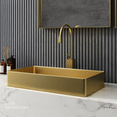 A modern bathroom showcasing the SUS304 Stainless Steel Rectangle Bathroom Sink in Brushed Gold by Mike Jake, set on a white marble countertop. Above the sink is a matching gold rectangular faucet. The wall behind features vertical gray stripes and a large rectangular mirror. To the left of the sink, two brown bottles and a reed diffuser complement other sleek bathroom faucets.