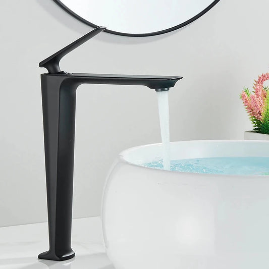 A Wasser™ Solid Brass Bathroom Sink Faucet by Rozin, featuring a sleek, angular design in modern black, dispenses water into a round, white vessel sink. In the background, a partial reflection is seen in a circular mirror, while a small plant adds a touch of greenery to the scene.