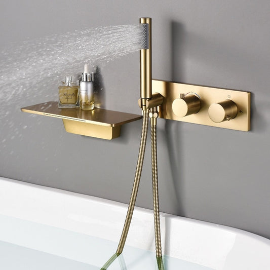 A Wasser™ Waterfall Bathroom Faucet with Handheld Shower Set by AllFixture, finished in modern gold, is mounted on a gray wall. The fixture includes an integrated shelf holding a few small bottles and features a control panel with two knobs, exemplifying the elegance of premium bathroom faucets. Below, a filled bathtub is visible.