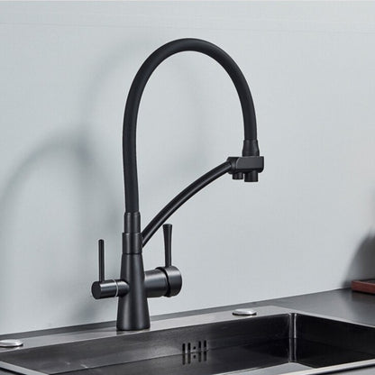 A person holds the spout of the Rozin Deck Mounted Brass Kitchen Faucet in black, directing a stream of water into a stainless steel sink filled with vegetables. The background features a sleek, minimalist kitchen with two white containers on the countertop.