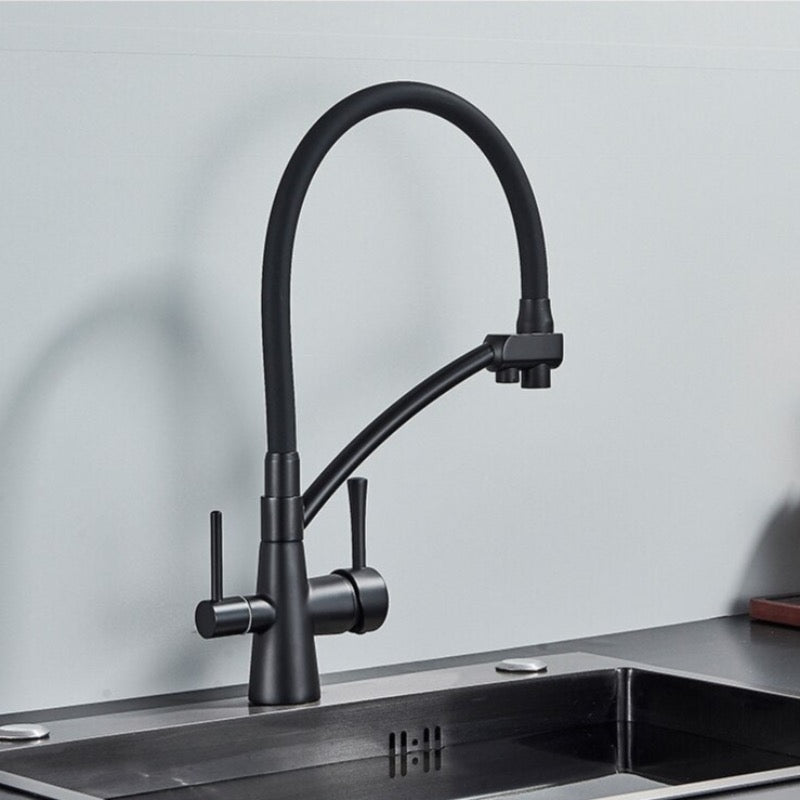 A person holds the spout of the Rozin Deck Mounted Brass Kitchen Faucet in black, directing a stream of water into a stainless steel sink filled with vegetables. The background features a sleek, minimalist kitchen with two white containers on the countertop.