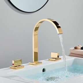 A modern bathroom sink features the Double Handle Deck Mounted Bathroom Faucet by AllFixture in polished gold, with a high-arching spout that gracefully dispenses water into the basin. The faucet has two square-shaped handles; one bears a red indicator for hot water, and the other displays a blue indicator for cold water. In the background, a round mirror can be seen.