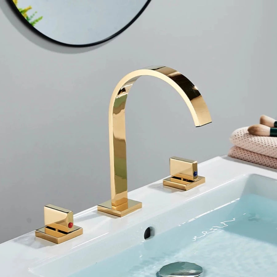 A modern bathroom sink features the Double Handle Deck Mounted Bathroom Faucet by AllFixture in polished gold, with a high-arching spout that gracefully dispenses water into the basin. The faucet has two square-shaped handles; one bears a red indicator for hot water, and the other displays a blue indicator for cold water. In the background, a round mirror can be seen.