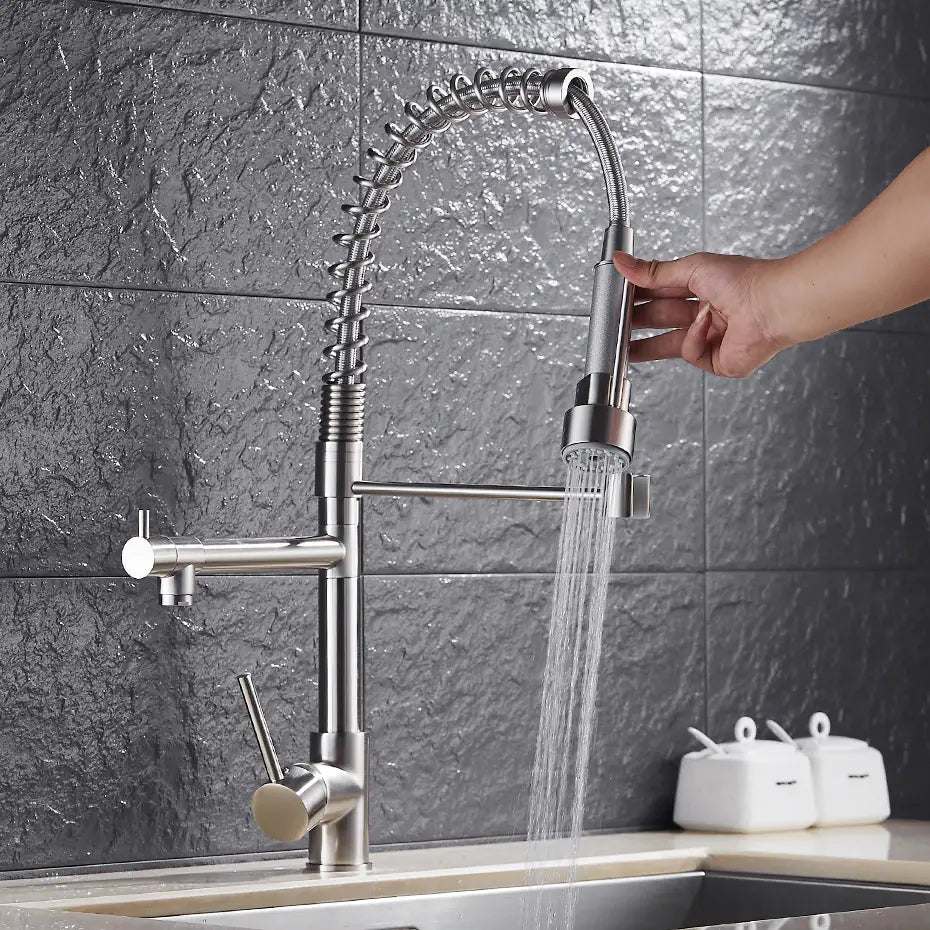 A Wanfan Pull Out Spring Spout Kitchen Faucet, featuring a modern stainless steel design and a flexible, spring-like hose, is shown in use. Water flows from the detachable spray head held by a person's hand over a sink with a gray tile backsplash. Soap dispensers are visible on the counter in the background.