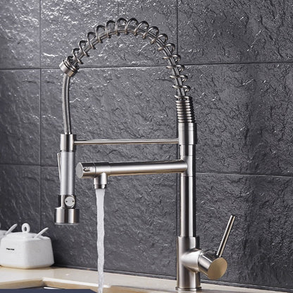 A Wanfan Pull Out Spring Spout Kitchen Faucet, featuring a modern stainless steel design and a flexible, spring-like hose, is shown in use. Water flows from the detachable spray head held by a person's hand over a sink with a gray tile backsplash. Soap dispensers are visible on the counter in the background.