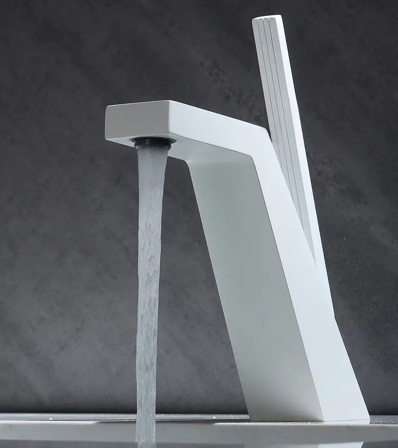 A modern, sleek Becola Single Handle Brass Bathroom Sink Faucet with an angular design pours water into a black sink. A blurred bottle and reed diffuser are in the background, set against a minimalist dark wall. The image is in black and white.