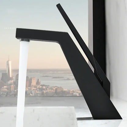 A modern, sleek Becola Single Handle Brass Bathroom Sink Faucet with an angular design pours water into a black sink. A blurred bottle and reed diffuser are in the background, set against a minimalist dark wall. The image is in black and white.