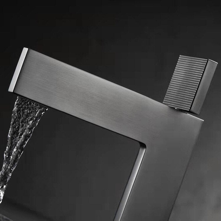 A modern, minimalist Single Handle Bathroom Basin Faucet by Mike Jake with a sleek, angular design is positioned above a textured, rectangular sink basin. The faucet and sink appear to be made of matte-finished materials, set against a dark, moody background.