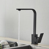 A contemporary Deck Mounted Matte Black Single Handle Kitchen Faucet by Rozin with a right-angled design is installed above the sink. Water is flowing from the faucet into the sink, with dishes stacked on a drying rack beside it. The background features a light-colored textured wall, reminiscent of sleek bathroom faucets found in modern homes.