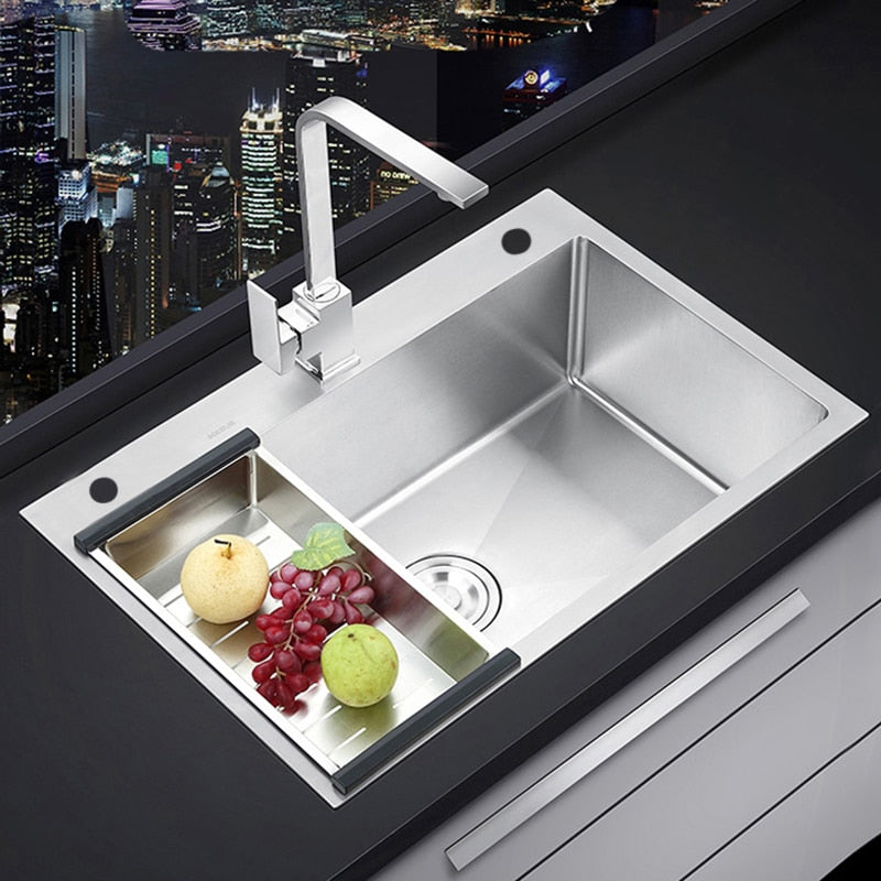 The AllFixture Single Bowl Handmade Stainless Steel Kitchen Sink showcases a modern and sleek design, complemented by a tall rectangular kitchen sink faucet. Inside the sink, there are green apples and a bunch of red grapes in a smaller compartment. The dark kitchen counter adds contrast, while the background reveals a nighttime cityscape.