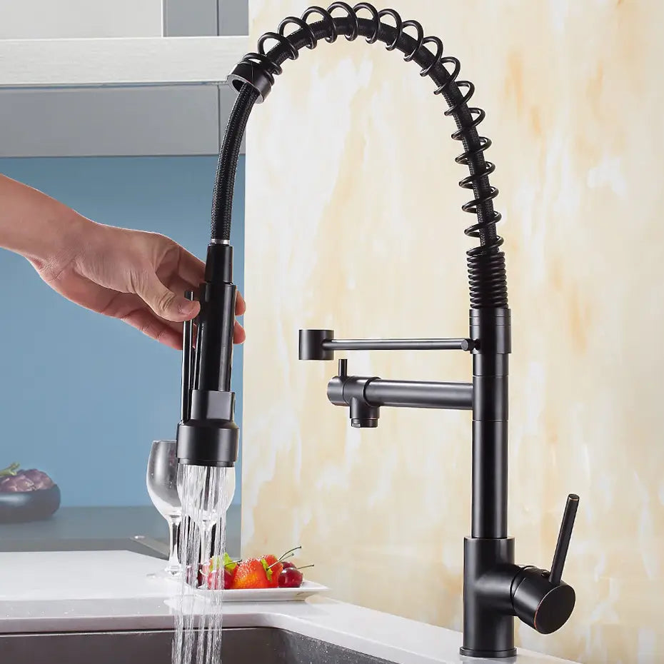 Hand holding the spray head of an allfixture Solid Brass Pull Out Spring Spout Kitchen Faucet with a matte black finish and flexible, coiled spring design, over a sink filled with strawberries. The faucet is turned on, and water is flowing from the spray head. Nearby, there is a wine glass and a cutting board with eggplants.