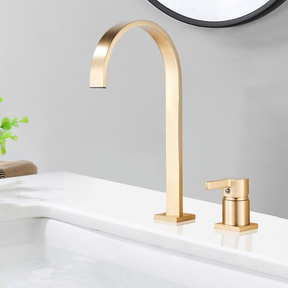 A brushed gold solid brass deck mounted bathroom sink faucet by allfixture showcases a modern high-arc spout and a single lever handle. Installed on a white countertop with a blurred plant and mirror in the background, this faucet creates a cohesive contemporary look, seamlessly complementing any kitchen sink faucet or shower head in matching finishes.