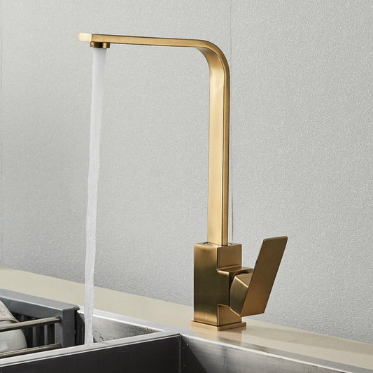 The Deck Mounted Single Handle Kitchen Faucet by Rozin, with its sleek and minimalist brushed gold finish, releases a steady stream of water into a stainless steel sink. It features a right-angled spout and an angular handle, set against a plain, light-colored wall.