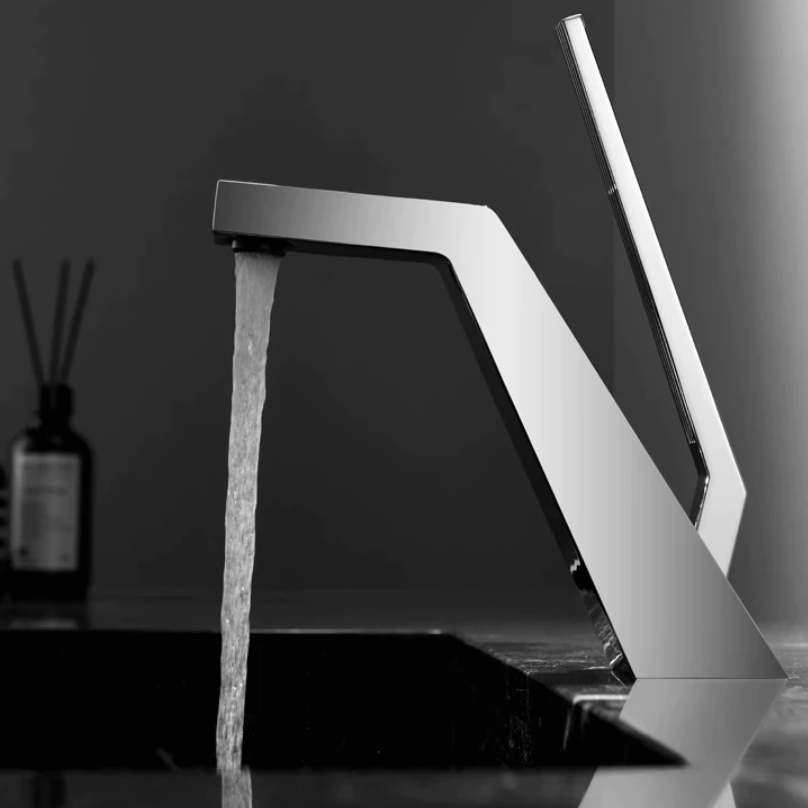 A modern, sleek Becola Single Handle Brass Bathroom Sink Faucet with an angular design pours water into a black sink. A blurred bottle and reed diffuser are in the background, set against a minimalist dark wall. The image is in black and white.