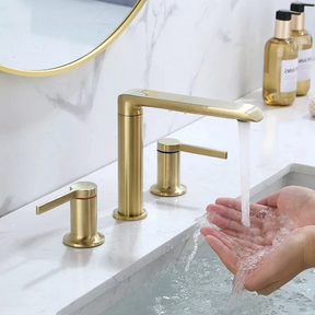 A modern bathroom sink features water flowing from the Solid Brass Deck Mounted Bathroom Sink Faucet by Morsale.com into a white basin. The countertop is crafted from white marble, and there are two matching chrome handles flanking the faucet. A round mirror and toiletries are visible in the background, creating an elegant ambiance reminiscent of a stylish kitchen faucet.