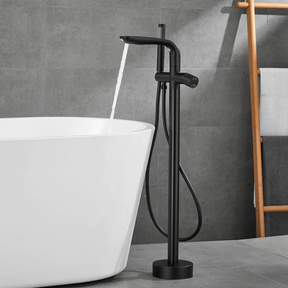 A modern bathroom features the Solid Brass Floor Bathtub Faucet Shower Mixer by Mike Jake, its gold tones pouring water into a freestanding white bathtub. The background is composed of gray tiles, and a wooden ladder shelf holding towels is partially visible. Adjacent to the setup, a sleek bathroom sink faucet enhances the contemporary design.