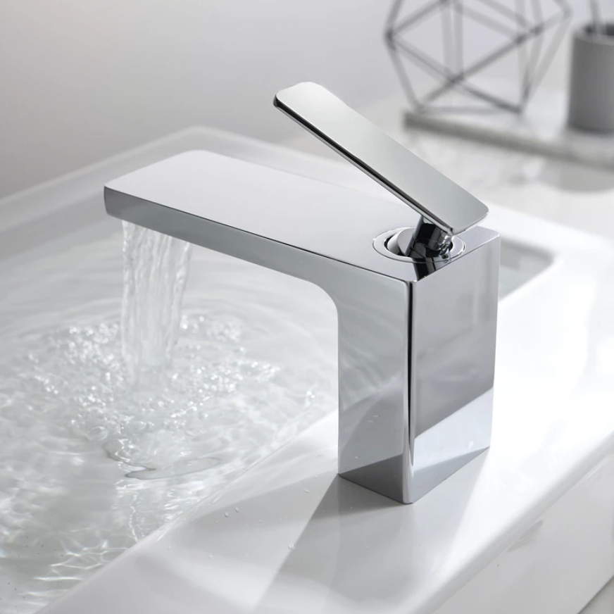 The Mike Jake Waterfall Style Solid Brass Bathroom Sink Faucet, with its modern rectangular design and angled lever handle, dispenses water into a white sink. In the background, a blurry geometric decoration and other bathroom essentials are visible. The faucet's minimalist and sleek aesthetic perfectly complements the setting.