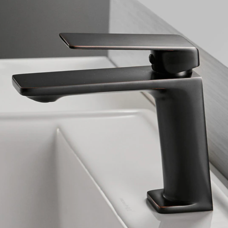 A sleek, modern Wanfan Single Handle Bathroom Sink Faucet in black, featuring clean lines and an angular design, is mounted on a white sink in the bathroom. The single lever handle is positioned over the base, creating a minimalist and contemporary look against a light grey background.