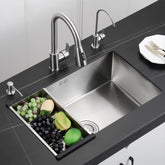 The AllFixture 304 Stainless Steel Single Bowl Undermount Kitchen Sink, with its sleek, modern design and stainless steel finish, includes a cutting board insert holding an assortment of fruits and vegetables such as grapes, an avocado, a lime, and an apple. Paired with a stylish kitchen sink faucet, the sink is elegantly set in a dark countertop with a plate and cutlery nearby.