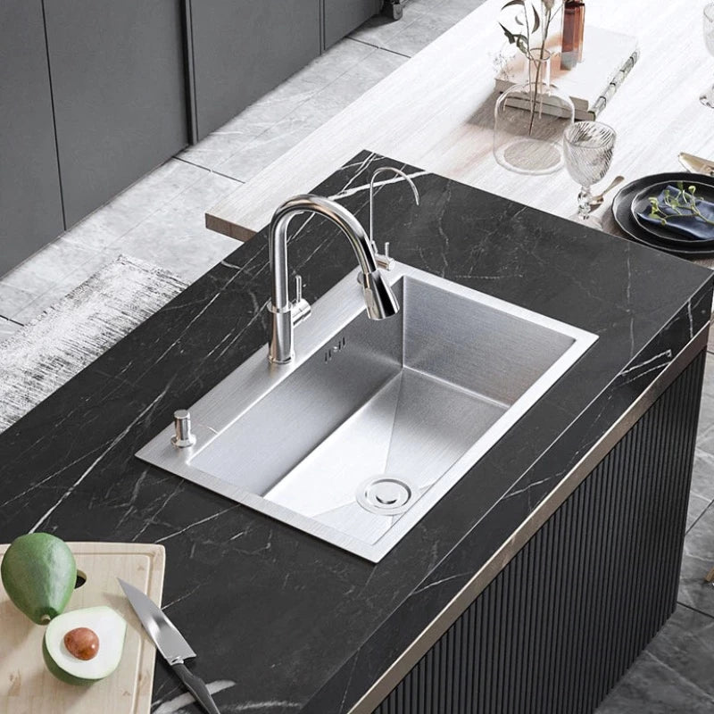 The AllFixture Single Bowl Handmade Stainless Steel Kitchen Sink showcases a modern and sleek design, complemented by a tall rectangular kitchen sink faucet. Inside the sink, there are green apples and a bunch of red grapes in a smaller compartment. The dark kitchen counter adds contrast, while the background reveals a nighttime cityscape.