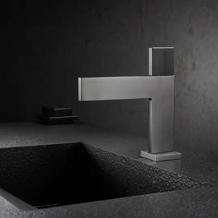 A modern, minimalist Single Handle Bathroom Basin Faucet by Mike Jake with a sleek, angular design is positioned above a textured, rectangular sink basin. The faucet and sink appear to be made of matte-finished materials, set against a dark, moody background.