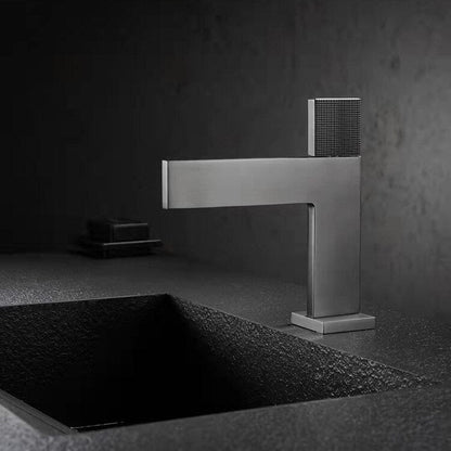 A modern, minimalist Single Handle Bathroom Basin Faucet by Mike Jake with a sleek, angular design is positioned above a textured, rectangular sink basin. The faucet and sink appear to be made of matte-finished materials, set against a dark, moody background.