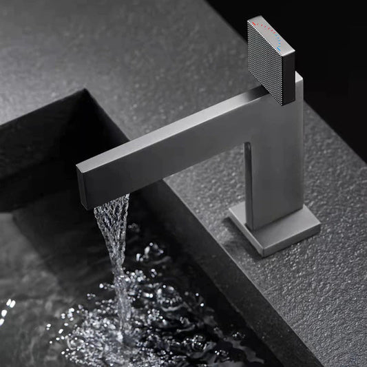 A modern, minimalist Single Handle Bathroom Basin Faucet by Mike Jake with a sleek, angular design is positioned above a textured, rectangular sink basin. The faucet and sink appear to be made of matte-finished materials, set against a dark, moody background.