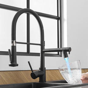 An AllFixture Single Handle Double Spout LED Kitchen Faucet with a sleek, modern black finish and a flexible spring neck dispenses water into a transparent glass. The faucet is mounted on a black countertop, set against wooden panels in the background. Natural light streams through a window framed in black in the background.