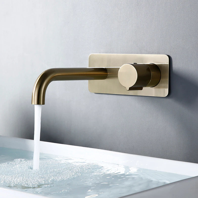 An elegant Wasser™ Wall Mounted Brass Bathroom Faucet by AllFixture is installed on a gray wall, gracefully pouring water into a pristine white sink below. The water runs smoothly, forming tiny bubbles in the sink, reminiscent of the gentle flow from a luxury shower faucet.