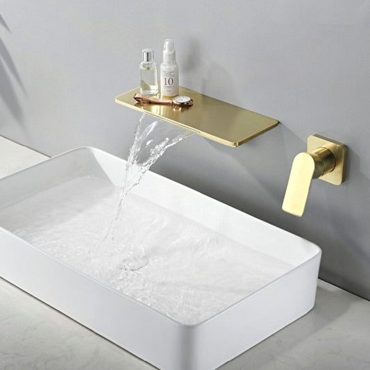 A modern bathroom sink setup featuring the AllFixture Solid Brass Wall Mounted Waterfall Sink Faucet extending horizontally from the wall in a gold finish. Water flows gracefully from the faucet into a rectangular white basin. Two small bottles are placed on the faucet shelf above the cascading water. The wall and countertop are decorated in muted tones.