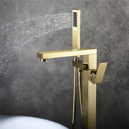 A modern brass Wasser™ Floor Mounted Bathtub Filler Shower Set by allfixture is installed against a gray wall. Water sprays horizontally from the handheld shower head attached to the fixture. The sleek, angular design and lever handle for controlling flow enhance its elegance. Below, the edge of a white bathtub is visible.