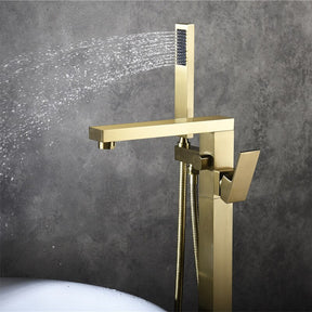 A modern brass Wasser™ Floor Mounted Bathtub Filler Shower Set by allfixture is installed against a gray wall. Water sprays horizontally from the handheld shower head attached to the fixture. The sleek, angular design and lever handle for controlling flow enhance its elegance. Below, the edge of a white bathtub is visible.