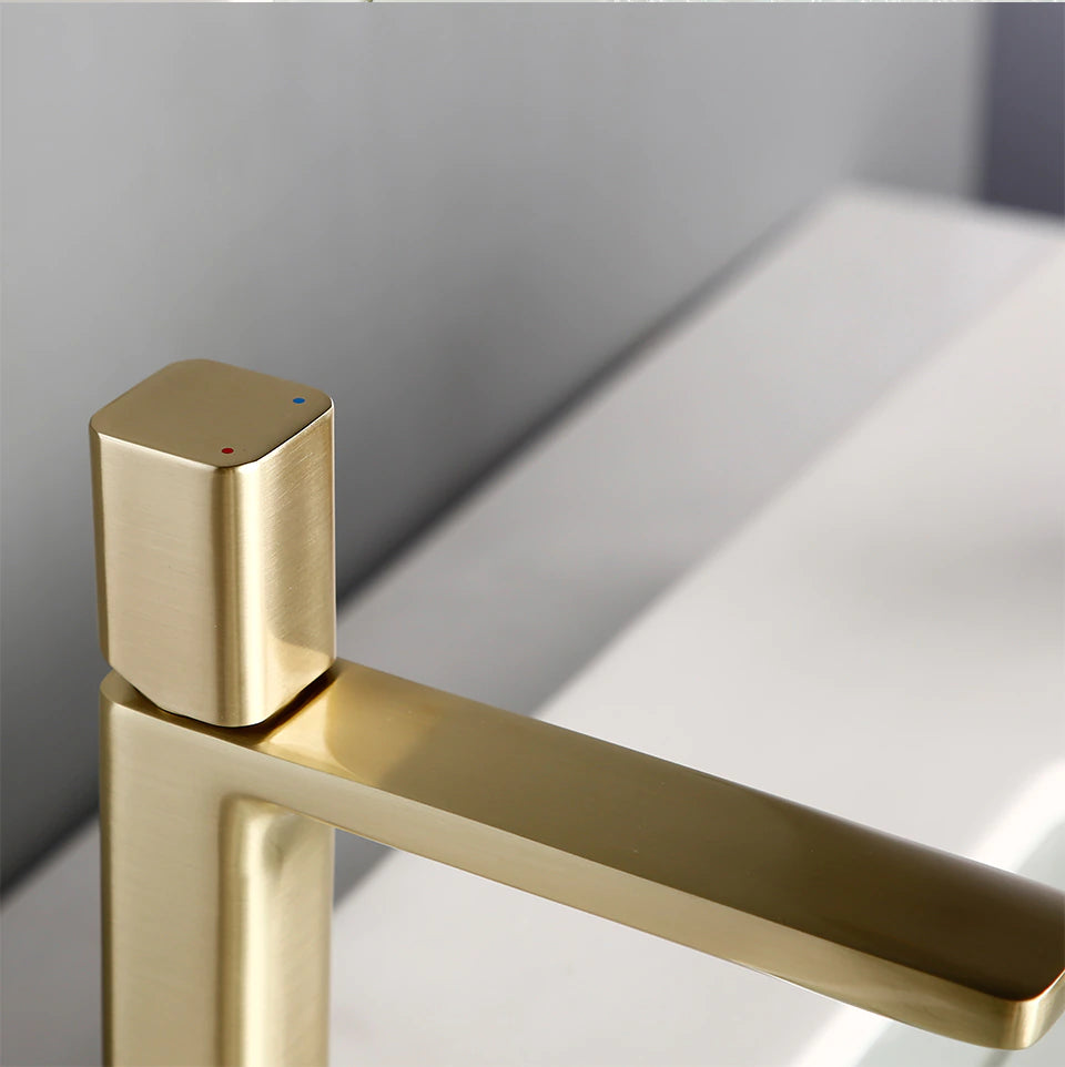 A sleek, modern Wasser™ Solid Brass Bathroom Sink Faucet from Morsale.com pours water into a rectangular white sink. The water flows smoothly, creating ripples in the filled basin. The background includes a gray wall and a corner of a wooden tray on the countertop.